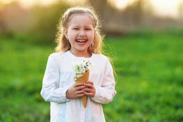 waffle cone filled with blooming cherry branches, held in the hands of a small smiling girl on a blurred natural background, at sunset. The concept of celebrating spring. Copy space. - Valokuva, kuva