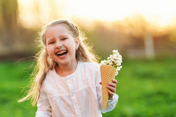 lovely little girl is laughing and holding the tree's spring flowers in a waffle cone in her hand. The concept is the beginning of a new, spring mood and childlike spontaneity. Warm sunset. - Zdjęcie, obraz