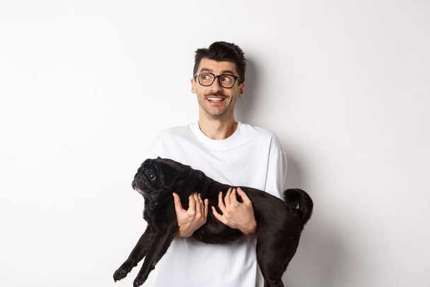Homme hipster excité avec son chien regardant à gauche le logo. Beau propriétaire d'animaux de compagnie portent carlin noir et regardant bannière promo, debout sur fond blanc - Photo, image