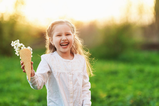 lovely little girl is laughing and holding the tree's spring flowers in a waffle cone in her hand. The concept is the beginning of a new, spring mood and childlike spontaneity. Warm sunset. - Foto, Imagen