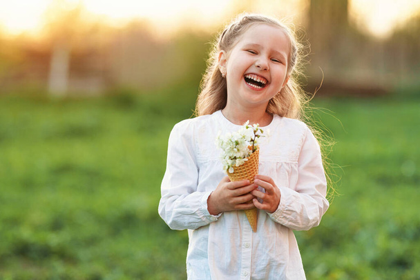 waffle cone filled with blooming cherry branches, held in the hands of a small smiling girl on a blurred natural background, at sunset. The concept of celebrating spring. Copy space. - Photo, image
