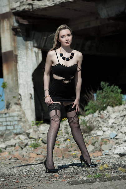 Beautiful sexy blonde girl in a black dress and stockings posing on a background of ruins - Photo, Image