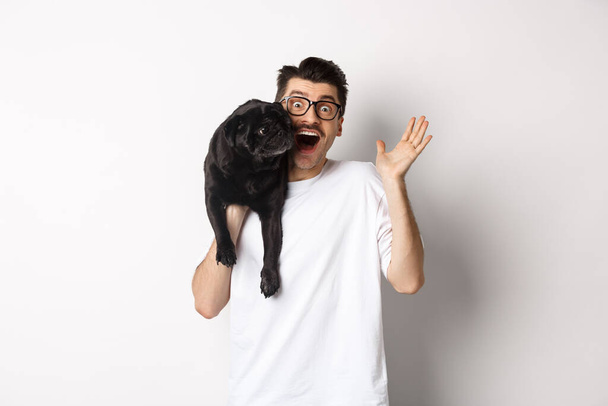 Gözlüklü yakışıklı genç adam siyah pug 'unu tutuyor ve el sallıyor, merhaba derken tek kollu köpek taşıyor, beyaz arka planda duruyor. - Fotoğraf, Görsel