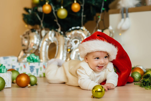 Cute smiling baby is lying under a festive Christmas tree and playing with gifts. Christmas and New Year celebrations - Foto, imagen