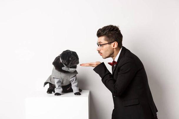 Pets, winter holidays and celebration concept. Handsome young man sending air kiss at cute black puppy wearing costume for New Year, owner standing in suit over white background - Photo, Image