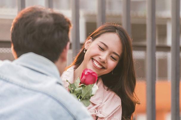 jong gelukkig paar liefde en romantisch op eerste date relatie. aziatische tiener vrouw verrassing en glimlachen naar vriendje geeft rode roos bloemen bij het diner in Valentijnsdag. koppel en geluk concept. - Foto, afbeelding