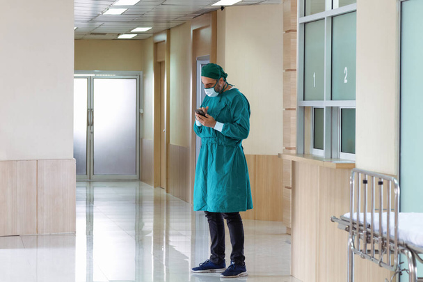 professional surgeon doctor in the gown and the protective mask is relaxing and using smartphone to calls family after success in serious case surgery in front of the operating room at the hospital. - Foto, immagini