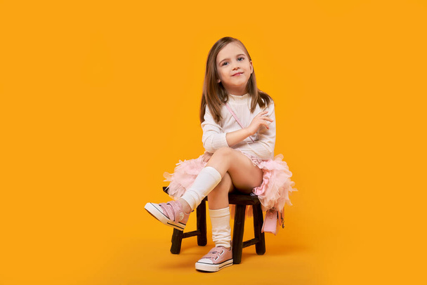 Young girl in tulle skirt and white pullover sitting on small wooden stool on bright yellow background - Foto, Bild