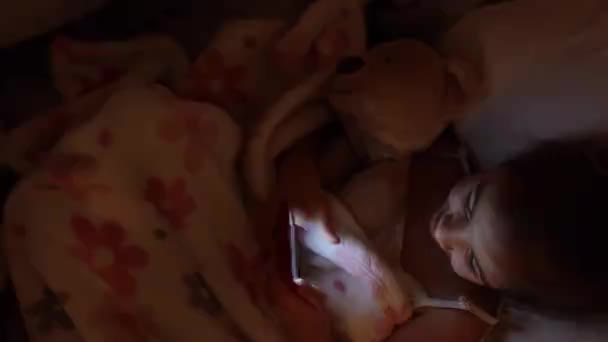 Dívka leží v posteli s měkkou hračkou a místo spánku používá smartphone. Koncept dětské závislosti na hrách. - Záběry, video