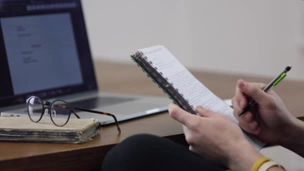 Een jongeman in het proces van werken op een laptop maakt notities en notities in een notebook. Studie en werkconcept. - Video