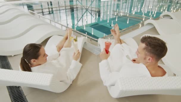 Imágenes en cámara lenta de la alegre pareja joven mirándose unos a otros tomando copas de cócteles, mientras se relaja con batas blancas en las tumbonas de la piscina termal techada - Metraje, vídeo