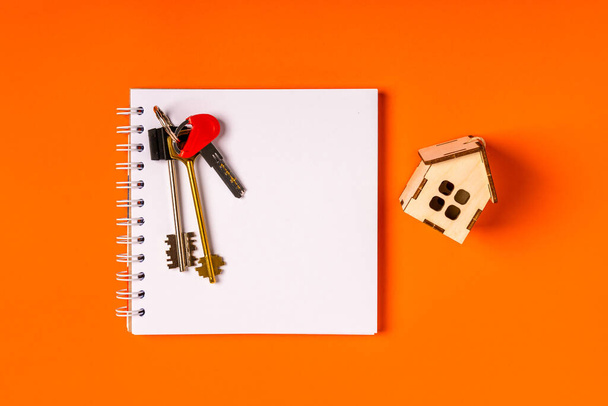 A empty notebook on a spiral, house keys and a wooden house on an orange background. Top view, real estate purchase concept - Photo, Image