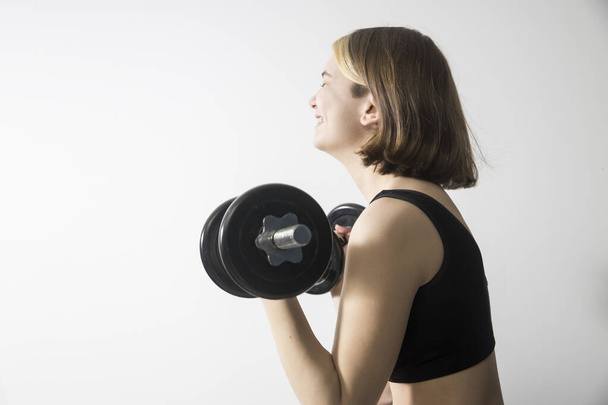 A nineteen-year-old smiling girl with a bob hairstyle and bleached strands in a gray short top holds a dumbbell on a white background. Sport and health concept. - Φωτογραφία, εικόνα