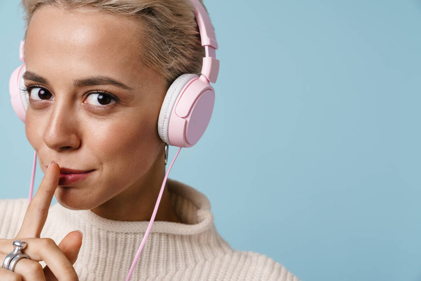 Pleased beautiful girl in headphones showing silence gesture isolated over blue background - Foto, Imagen