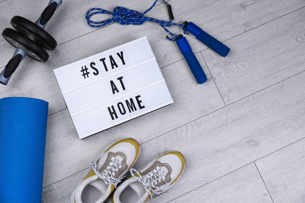 Sport equipment and lightbox with hashtag STAY AT HOME on floor, flat lay. Message to promote self-isolation during covid19 pandemic - Fotoğraf, Görsel