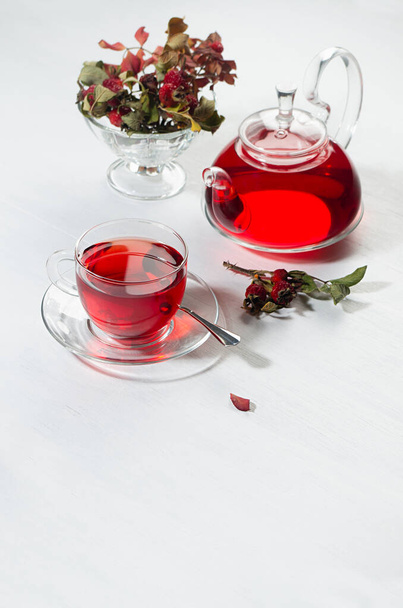 Autumn teatime with red herbal tea in transparent tea pot, cup with hawthorn berries, rose hip, dry leaves in sunlight on white wood table, vertical. - Фото, зображення