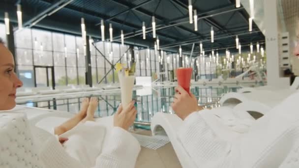 Zoom-in slow-motion of young caucasian couple wearing white bathrobes chilling on loungers by roofed thermal pool clinking with glasses of refreshing cocktails looking at each other - Footage, Video