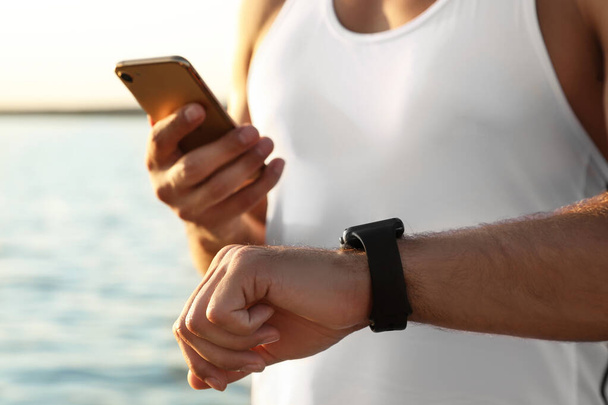Man with fitness tracker and smartphone checking results after training near river, closeup - Fotoğraf, Görsel