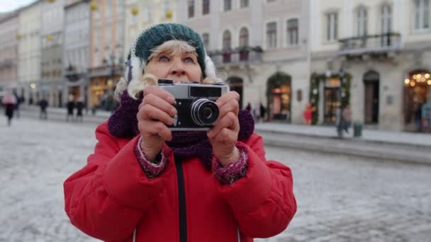 Senior vieja turista tomando fotos con cámara fotográfica, utilizando dispositivo retro en el centro de la ciudad de invierno - Metraje, vídeo