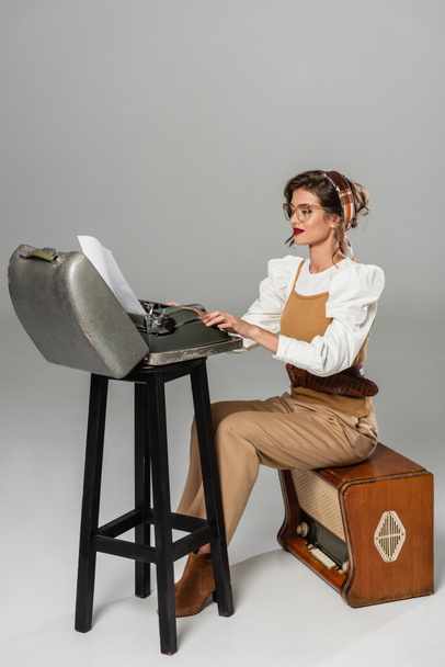 secretary in retro clothes sitting on radio receiver and typing on vintage typewriter on grey - Fotoğraf, Görsel