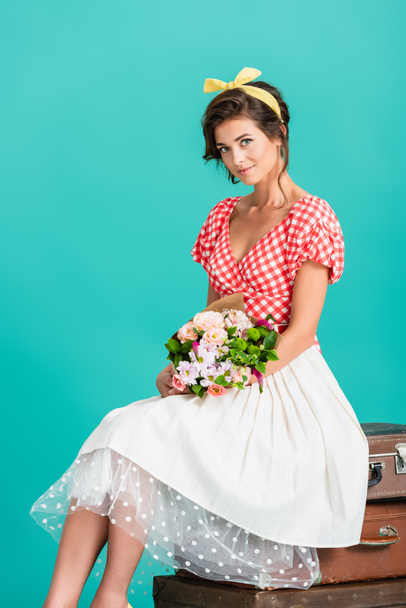 smiling woman in retro clothes sitting on vintage suitcases with flowers isolated on turquoise - Foto, imagen