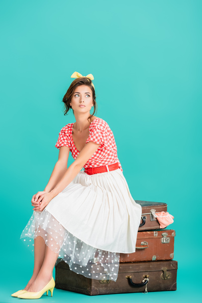 pretty pin up woman sitting on vintage suitcases and looking away on turquoise - Fotoğraf, Görsel