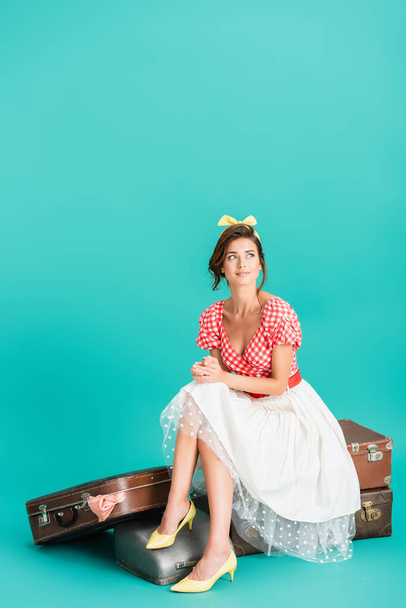 smiling pin up woman looking away while sitting on vintage suitcases on turquoise - Fotó, kép