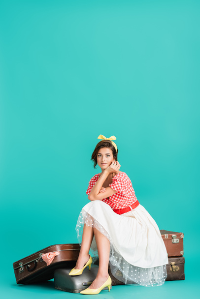 pretty pin up woman sitting on vintage suitcases and looking at camera on turquoise - Foto, Imagem