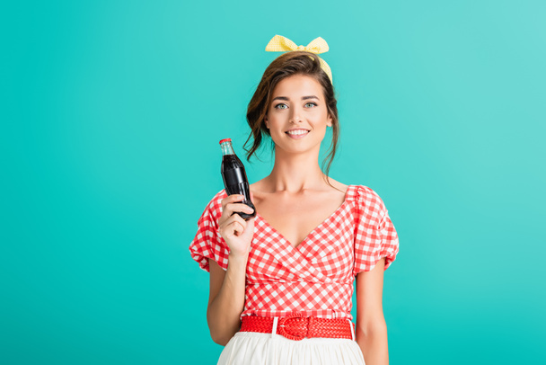 mujer feliz en ropa retro de moda sonriendo a la cámara mientras sostiene la botella de refresco aislado en turquesa - Foto, imagen