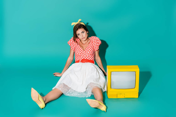 joven pin up mujer mirando a la cámara mientras está sentado en el suelo cerca de amarillo vintage tv en turquesa - Foto, Imagen