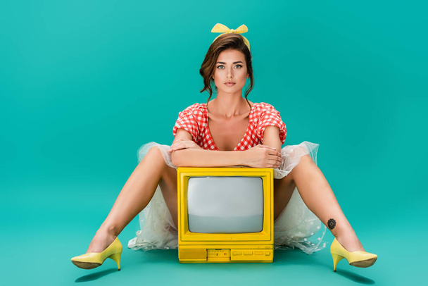 joven pin up mujer mirando a la cámara mientras está sentado cerca de brillante amarillo vintage tv en turquesa - Foto, Imagen