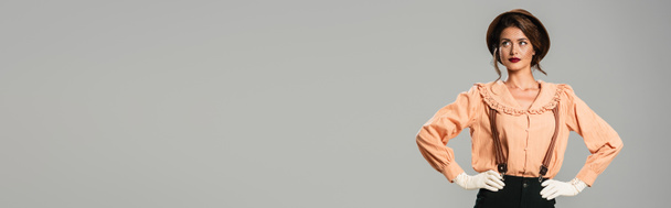 ładna młoda kobieta w modne ubrania retro i rękawiczki pozowanie z rękami na biodrach odizolowanych na szary, sztandar - Zdjęcie, obraz