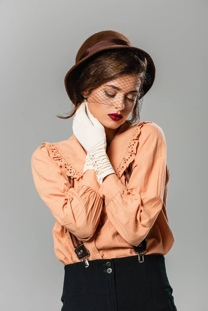 junge Frau mit Vintage-Mütze und Handschuhen, die den Hals berühren, während sie mit geschlossenen Augen auf grau isoliert posiert - Foto, Bild