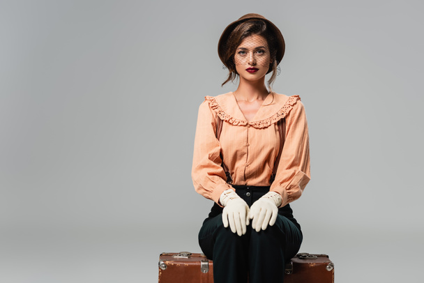 молодая женщина в шляпе и модной ретро-одежде сидит на винтажном чемодане на сером - Фото, изображение
