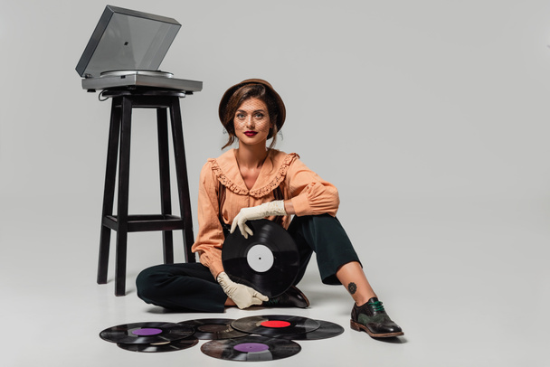 junge Frau in Retro-Klamotten blickt in die Kamera, während sie auf grauen Plattenspielern und Schallplatten sitzt - Foto, Bild