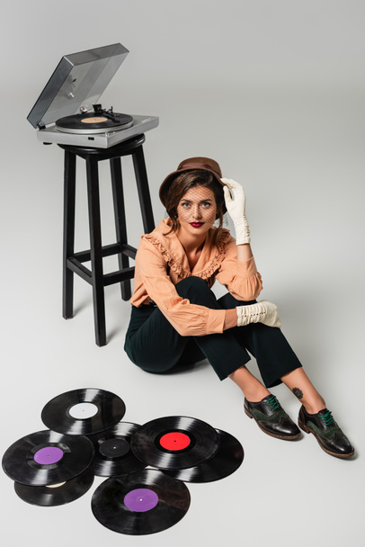 Hochwinkelaufnahme einer stilvollen Frau, die ihren Hut berührt, während sie in der Nähe von Schallplatten und Plattenspieler auf grau sitzt - Foto, Bild