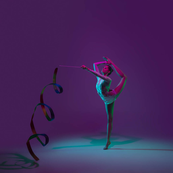 Young female athlete, rhythmic gymnastics artist on purple background with neon light. Beautiful girl practicing with equipment. Grace in performance. - Foto, immagini