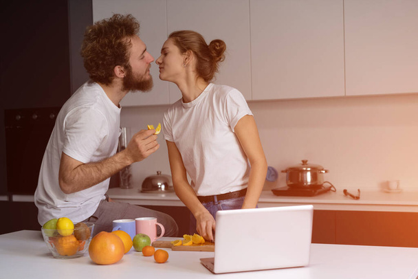 Beautiful young couple kissing feeding each other making fun at modern kitchen and smiling while cooking at home. Pretty girl feeds or nursing her boyfriend - Фото, зображення