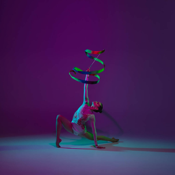 Young female athlete, rhythmic gymnastics artist on purple background with neon light. Beautiful girl practicing with equipment. Grace in performance. - Foto, imagen