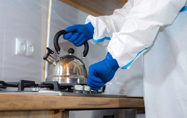 Arzt in medizinischem Schutzanzug, Schutzbrille, Maske und Handschuhen bereitet Tee in der Küche zu. Teekanne auf dem Gasherd. Schutz vor Virusepidemie. Coronavirus (COVID-19). Gesundheitskonzept. - Foto, Bild