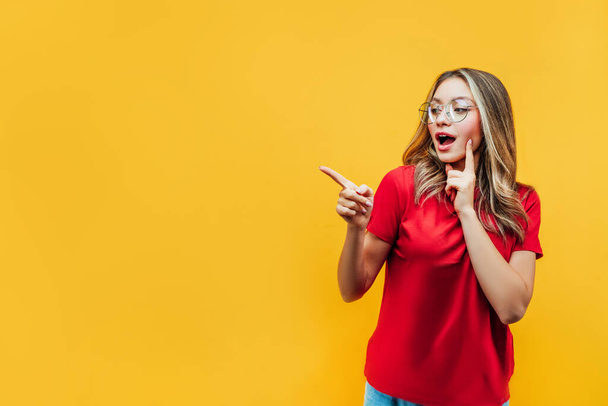 Pretty girl in a red T-shirt and transparent glasses on a yellow background shows his hand in place for text and looks surprised. - Valokuva, kuva