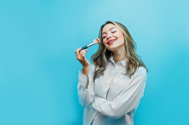 Beautiful girl on a blue background with a bone for makeup in hand, charmingly smiling. Copy space. - Fotó, kép