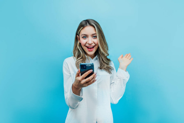 Charming girl on a blue background holds a phone in his hand and looks at the camera in surprise. Place for text. - Fotó, kép