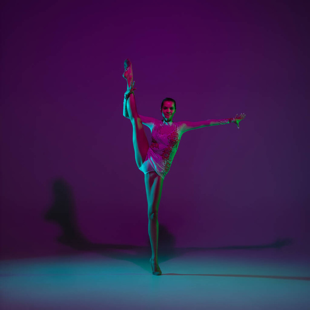 Young female athlete, rhythmic gymnastics artist on purple background with neon light. Beautiful girl practicing with equipment. Grace in performance. - Foto, Imagen