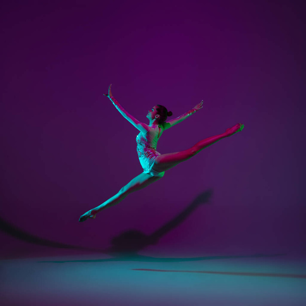 Young female athlete, rhythmic gymnastics artist on purple background with neon light. Beautiful girl practicing with equipment. Grace in performance. - Valokuva, kuva