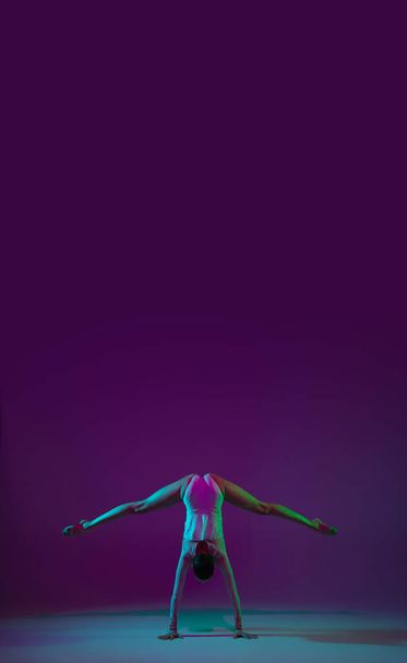 Young female athlete, rhythmic gymnastics artist on purple background with neon light. Beautiful girl practicing with equipment. Grace in performance. - Foto, afbeelding