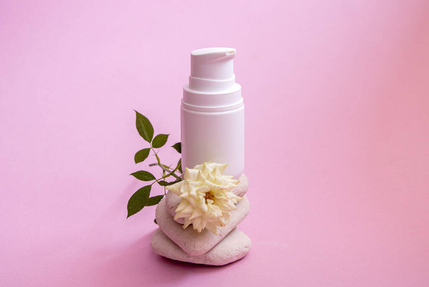 a white plastic jar with a dispenser and a gentle caring organic hand and body cream, on smooth textured rounded stones on a pink background with a small room living white rose. - Photo, image