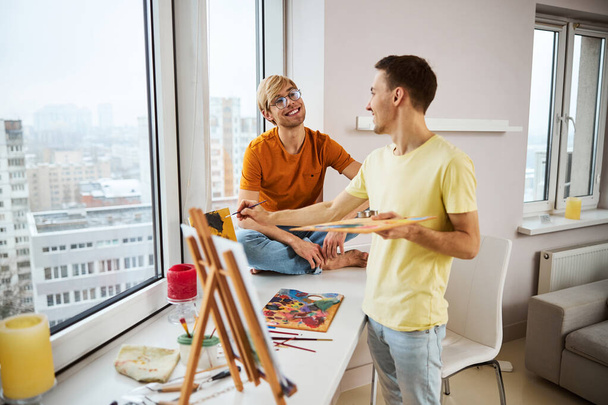Homme joyeux peinture tableau et bavarder avec petit ami à la maison - Photo, image