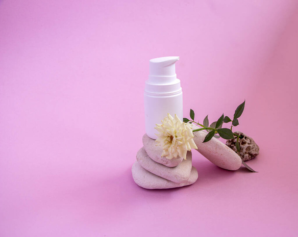 a white plastic jar with a dispenser and a gentle caring organic hand and body cream, on smooth textured rounded stones on a pink background with a small room living white rose. - Fotografie, Obrázek