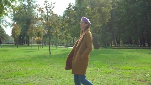 jovem mulher em boina e casaco andando com as mãos em bolsos no parque de outono - Filmagem, Vídeo